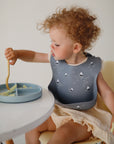 A toddler with curly hair wearing a blue sailboat bib sits at a table, using a fork to eat sliced fruit from the mushie Silicone Suction Plate, designed for easy baby feeding.