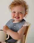 A smiling toddler with curly blond hair sits on a cream chair, holding a mushie Snack Cup and wearing a gray bib with sailboat designs against a plain background.