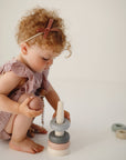 A young child with curly hair in a mauve outfit stacks mushie’s non-toxic Stacking Rings Toy on a wooden rod, while extra pastel-colored rings are scattered nearby on the white floor.