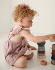 A young child with curly hair in a pink romper and headband sits on the floor stacking mushie Stacking Cups Toy, colorful pieces inspired by Danish Hygge design, building towers on a white background.