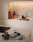 Modern kitchen with white cabinets and marble surfaces, featuring cutting boards and utensils by the sink. In the foreground, stacked plates, utensils, and a mushie Silicone Feeding Spoon with silicone bibs rest on a marble island.