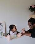 An adult feeds a smiling baby at a table with a bowl, cup, and sippy cup on a mushie Silicone Placemat. A plant and framed artwork in the background add to the warm, homey feel.