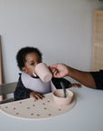 A baby sits at a table with food while being helped to drink from a cup. A mushie Silicone Feeding Spoon rests nearby as the baby looks up, ready for their next bite.