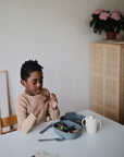 A young boy in a beige sweater sits at a white table with a mushie Lunchbox, featuring non-toxic plastic and built-in compartments for fruit, a sippy cup, and cutlery. A wooden cabinet with pink flowers and a framed print are in the background.