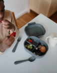 A child sits at a white table eating a strawberry from a mushie Lunchbox with built-in compartments filled with grapes, blueberries, apple slices, and more strawberries. A cup, fork, and spoon are also on the table.