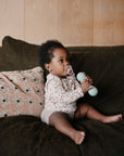 A baby with a pacifier sits on a dark green couch, holding the mushie Silicone Baby Rattle Toy made from food-grade silicone. A patterned pillow is beside the baby, and a wooden wall forms the background.