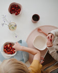 Two young children sit at a table with strawberries and cherries, using mushie Silicone Placemats made from food-grade silicone to keep snacks tidy as one child reaches for cherries. A cup, spoon, and a small vase with flowers are also on the table.