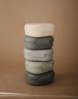 Five mushie Lunchboxes in cream, dark gray, light gray, sage green, and charcoal—each rectangular with built-in compartments and made from non-toxic plastic—are stacked vertically against a plain brown background.