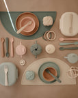 A flat lay of mushie Lunchbox feeding sets—pastel bowls, plates, utensils, sippy cups, snack containers with compartments, and textured teething toys—all made from non-toxic plastic and neatly arranged on beige placemats.