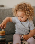 A young child in a light blue shirt and light purple pants sits on a couch, stacking mushie Stacking Rings Toy—made of non-toxic, colorful rings—while holding a pacifier. The child has curly blonde hair.