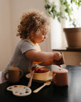 A curly-haired toddler sits at a table with strawberries, pouring a drink from a spill-proof cup. On the table is a mushie Snack Cup, pink mug, spoon, and round paint palette. A potted plant can be seen in the background.