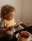 A young child with curly hair and a bib sits at a table, holding a BPA-free mushie Round Dinnerware Bowl. In front are a mug and baby bowls filled with strawberries and banana slices, creating a softly lit, cozy scene.