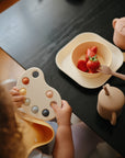 A young child sits at a dark table holding a sensory fidget toy. On the table are mushie Round Dinnerware Bowls with strawberries, a fork, and a beige cup with straw plus matching lidded container from the set.