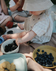 A toddler wearing a sun hat and bib sits on a blanket outdoors, reaching for blackberries in a mushie Square Dinnerware Bowl. Nearby are bowls of blueberries and cookies, with other children partially visible in the background.