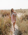 A woman walks barefoot along a sandy path near the beach, carrying a baby wrapped in a mushie Organic Cotton Muslin Swaddle Blanket on a sunny day.