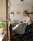 A woman wearing a hat smiles as she adjusts the mushie Multi-Use Cover in green on a stroller, surrounded by plants and shelves with ceramic dishes and jars in a sunlit, cozy room.