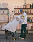 A person in a checkered jacket and blue cap stands beside a stroller featuring the mushie Multi-Use Cover, holding a pot of green plants. Behind them, shelves with plant pots and watering cans line a light-colored wall.