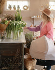 A woman wearing a pink sweater and brown hat holds a baby car seat with the mushie Multi-Use Cover while choosing white tulips from glass vases in a flower shop.