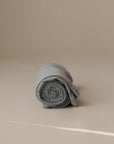 The mushie Multi-Use Cover, neatly rolled in light gray, sits on a smooth beige surface with a plain beige background.