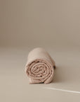 A neatly rolled mushie Multi-Use Cover in beige, ideal as a car seat or nursing cover, rests on a smooth matching surface with a minimal background.