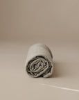 A neatly rolled, light gray mushie Multi-Use Cover is displayed on a smooth beige surface with a neutral beige background.