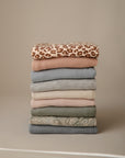A neatly stacked pile of neutral-toned clothes features mushie's Multi-Use Cover, with a brown and tan leopard print on top and a subtle paisley pattern beneath—ideal as a stylish nursing or multi-use cover.