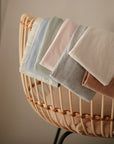 Soft pastel baby blankets drape over a wicker bassinet with a peach cushion, set against a neutral backdrop—ideal when paired with the mushie Multi-Use Cover for extra versatility.