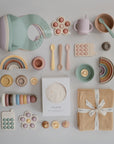 A flat lay displays pastel baby items, including mushie’s Stacking Rings Toy, bibs, spoons, toys, cups, plates, cloths, silicone rainbows, and a boxed muslin cloth—all neatly arranged on a light surface.