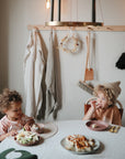 Two young children sit at a round table eating snacks on mushie Silicone Placemats. Plates of fruit and bread rest on the table, while clothes, utensils hang on hooks, and a decorative wreath is displayed in the background.