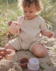 A toddler in a beige romper sits on sandy grass, holding a toy teapot and the mushie Snack Cup. Stacked toy cups are nearby, and another hand reaches into the frame—ideal for playtime with toddlers.