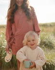 A woman in a pink dress stands behind a smiling blond child holding a light blue mushie Snack Cup. They are outdoors in a grassy, sunlit field, and the woman holds a small artist's palette.