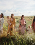 Four women stroll through a sunny field with toddlers, all in loose earth-toned outfits. One child carries the mushie Snack Cup, a spill-proof silicone container, as tall grass and open sky create a peaceful setting.