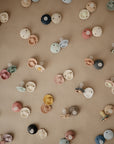 FRIGG Daisy Silicone Pacifier 2-Pack by FRIGG features colorful, daisy-shaped shields and medical-grade silicone nipples, displayed scattered across a neutral beige surface.