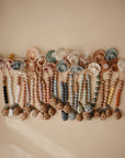 A row of FRIGG Moon Silicone Pacifier 2-Packs with beaded clips, arranged on a beige surface. The beads combine wood and medical grade silicone in soft pastel and neutral colors—ideal baby essentials from FRIGG.