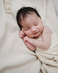 A newborn with dark hair sleeps soundly wrapped in a mushie Organic Cotton Muslin Swaddle Blanket featuring small animal prints, resting against someone in a ribbed, light garment. A pacifier clip lies nearby.