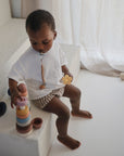 A toddler sits barefoot in a bright white room, playing with the mushie Stacking Rings Toy. They're wearing a white shirt and striped shorts, and a pacifier clip hangs from their shirt.