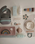 Flat lay of pastel baby feeding essentials, including mushie Stacking Rings Toy, non-toxic silicone bibs, spoons, bowls, cups, divided plate, sippy cup, pacifier clips, washcloths, and bottles of lotion and shampoo.