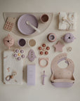 A flat lay of pastel mushie baby items—including a silicone bib, plates, bowls, toys, pacifier, teethers, cloths, and the mushie Snack Cup for toddlers—are neatly arranged on a cream background.