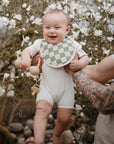 A smiling baby in a mushie Muslin Bib and white onesie is held by a tattooed adult in front of a blossoming tree with white flowers.