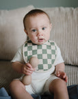 A baby wearing a mushie Muslin Bib in green and white check sits on a beige couch, holding a beige rattle and looking toward the camera.