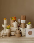 A beige and brown mushie Lunchbox made from non-toxic plastic is arranged neatly against a neutral background, alongside stacking cups, sippy cups, containers, and vases with yellow-orange flowers.