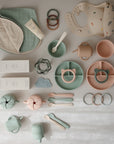 A flat lay of mushie’s pastel-colored baby feeding items—including a silicone Snack Cup, spill-proof plates, bowls, cups, bibs, teethers, and cloths—are neatly arranged for toddler mealtimes on a light surface.