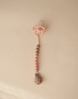 The FRIGG Andersen Fairytale Silicone Pacifier 2-Pack by FRIGG, made in Denmark, features a pink silicone pacifier attached to a beaded silicone and wooden clip, shown on a light beige background.