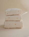 The mushie Extra Soft Bassinet Muslin Crib Sheet features a cream color with yellow sun patterns, stacked on a beige background—perfect as breathable baby bedding or a cozy crib sheet.