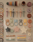 A neatly arranged collection of pastel mushie baby items—including stacking cups, teethers, spoons, pacifiers, toy rings, lids, and the Flower Training Toothbrush—are organized in rows on a light surface.