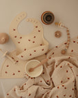 A beige baby bib and placemat with fox prints, mushie Organic Cotton Muslin Swaddle Blanket, a white bowl with wooden spoon, beige sippy cup, teether, pacifier clip, and stacked cups are arranged on a soft cream background.