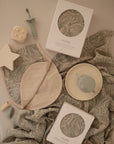 Baby products are arranged on a paisley-patterned cloth, including two boxed bibs, a mushie Organic Cotton Muslin Swaddle Blanket, a silicone sippy cup on a plate, soft star and cube teethers, and a pacifier clip in soft neutral colors.