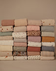 A tidy stack of mushie Organic Cotton Muslin Swaddle Blankets arranged in three rows, showcasing pastel stripes, florals, animals, and abstract patterns, all set against a plain beige backdrop.