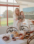 A woman holding a baby stands by a dining table set with plates, cups, and mushie Silicone Placemats, in a sunlit room featuring large windows and autumn-themed centerpieces.