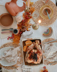 A festive table set for a meal features roast chicken, plates, cutlery, candles, glasses, autumn leaves, berries, and a soft mushie Silicone Feeding Spoon on a white tablecloth with woven placemats and beige napkins.